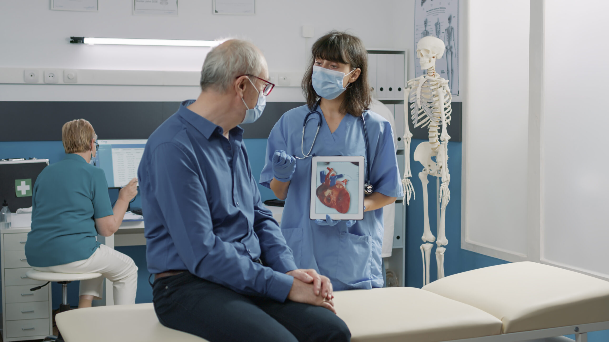 Female assistant holding tablet with cardiovascular system illustration, explaining heart condition to retired person during covid 19 pandemic. Nurse showing cardiology diagnosis on gadget.