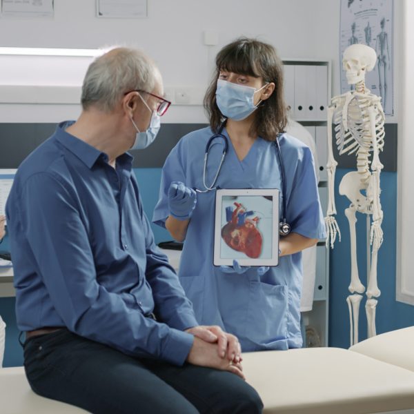 Female assistant holding tablet with cardiovascular system illustration, explaining heart condition to retired person during covid 19 pandemic. Nurse showing cardiology diagnosis on gadget.
