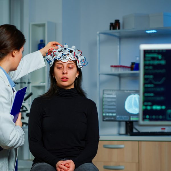 Neurological doctor cheking eeg headset explaining to patient diagnosis of disease and treatment for nervous system. Scientist researcher analysing brain scan and tomography on monitor in lab