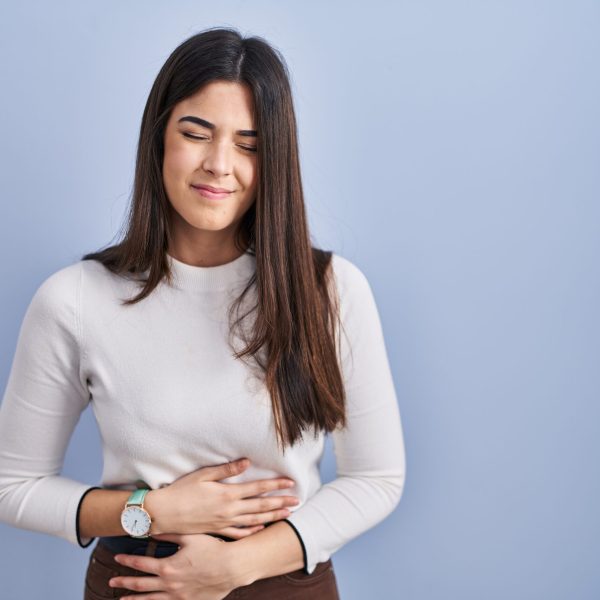 Young brunette woman standing over blue background with hand on stomach because indigestion, painful illness feeling unwell. ache concept.